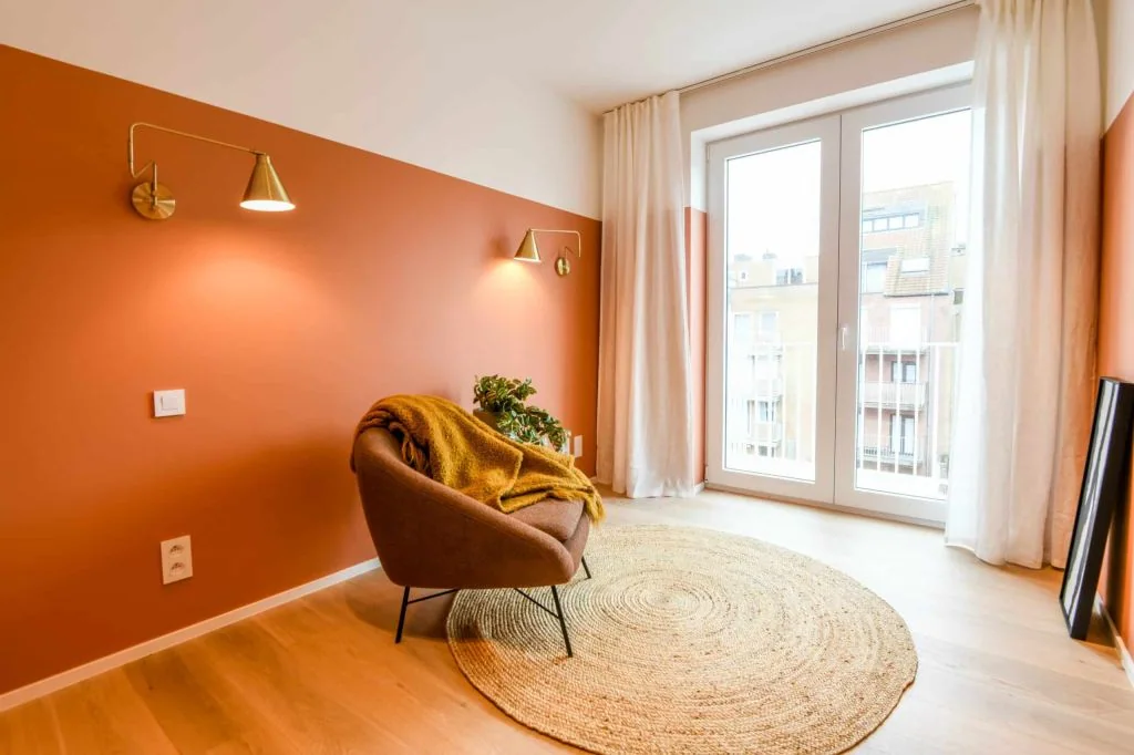 Mur d’accent peint en orange avec des appliques murales et un fauteuil près d’une grande fenêtre.